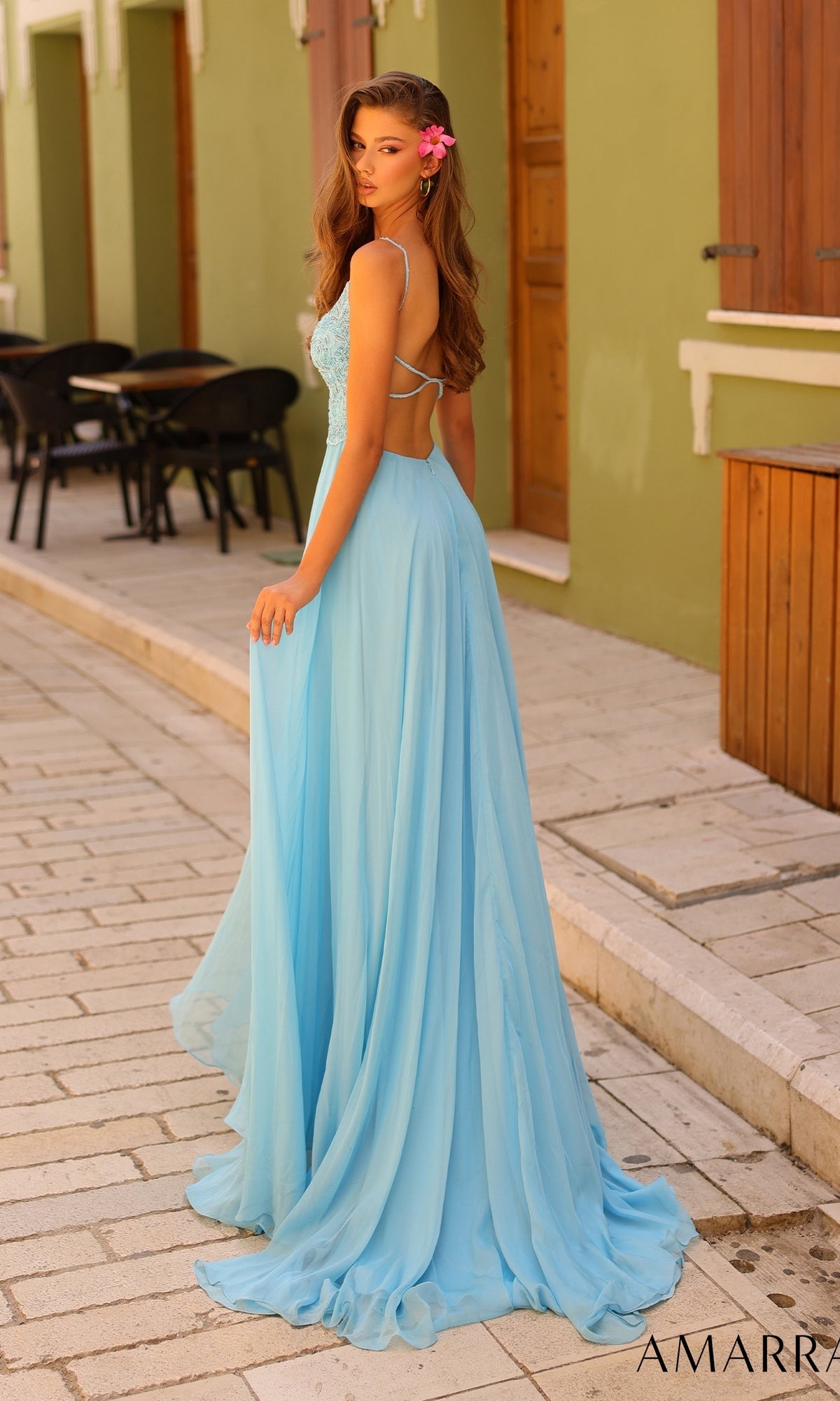 A woman in the Amarra Open-Back Long A-Line Prom Dress 94008, light blue in color, with a pink flower in her hair, stands on a stone walkway before green buildings.